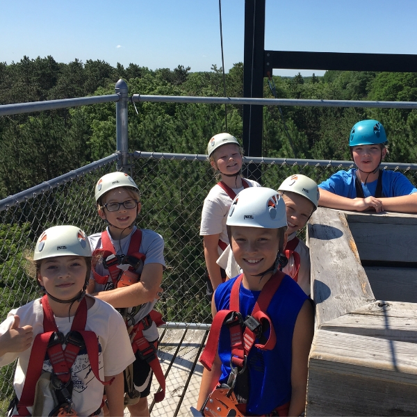 Smiling children wear helmets and harnesses, stand at top of zipline platform