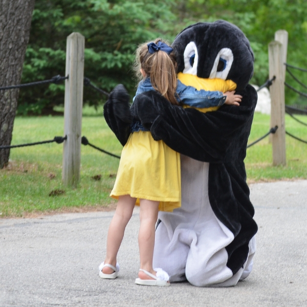 Person wearing penguin costume hugs girl in yellow dress