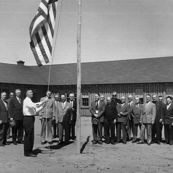 1950: Men stand in front of Reforestation Camp building raising a flag
